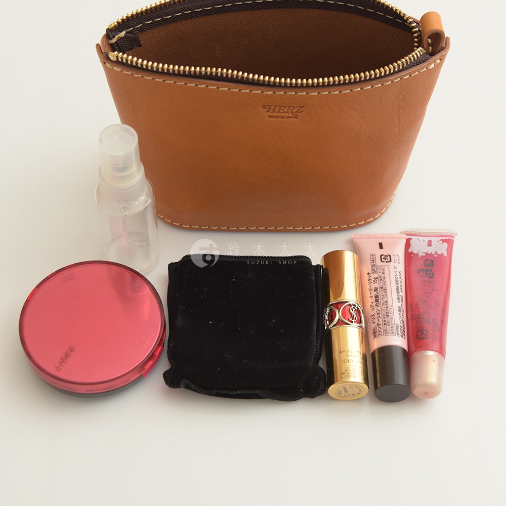 棕色皮革職人手作三角收納包，拉鍊開啟，內有空間。包包下方擺放著透明噴瓶、紅色圓形粉餅盒、黑色絨布收納袋、金色唇膏及兩支粉紅色護唇膏。