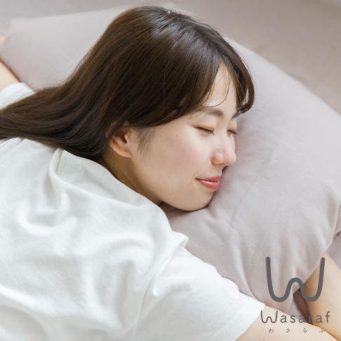 一位女子側臥在淺紫色雙人床包上，頭枕著同色系枕頭，臉部表情安詳。女子穿著白色短袖上衣，長髮散落在肩上。