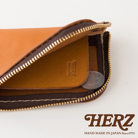 特寫視角展示日本HERZ職人手作皮革迷你錢包內部細節，錢包採用棕色皮革搭配深棕色拉鍊，皮革表面壓印品牌標誌與產地資訊，內部設有夾層並放入一枚硬幣作為比例參考，整體呈現簡約精緻的質感。