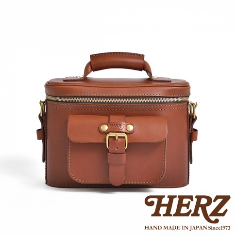 日本HERZ職人手作皮革厚實相機包，棕色皮革材質，正面有翻蓋口袋和黃銅扣帶，頂部有手提把，白色背景。