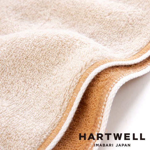 這張照片呈現了 Hartwell 今治午睡貓咪雙色毛巾的細部特寫，材質呈現柔軟蓬鬆的米白色毛圈質感，邊緣處則有對比鮮明的金棕色裝飾滾邊，畫面下方印有品牌名稱與產地標示。