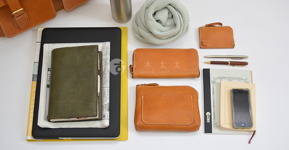 皮革旅行包 A-102，包含一個橄欖綠色皮革筆記本、一個卡其色長夾、一個卡其色零錢包、兩個鋼筆、一個手機，以及一個淺灰色針織圍巾。