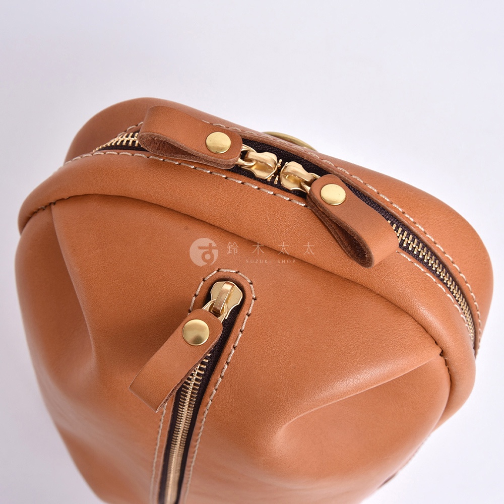 一個駝色皮革斜背包的特寫，上面有兩個金色的拉鍊和細節，右側印有日文和英文的 SUZUKI SHOP 標誌