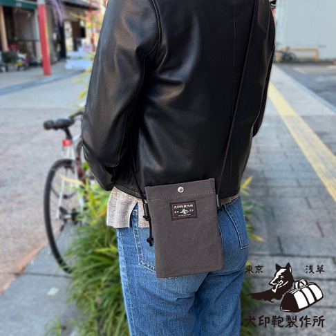 一名男子穿著黑色皮夾克和牛仔褲，背著灰色的帆布手機包，包上有犬印鞄製作所的標誌