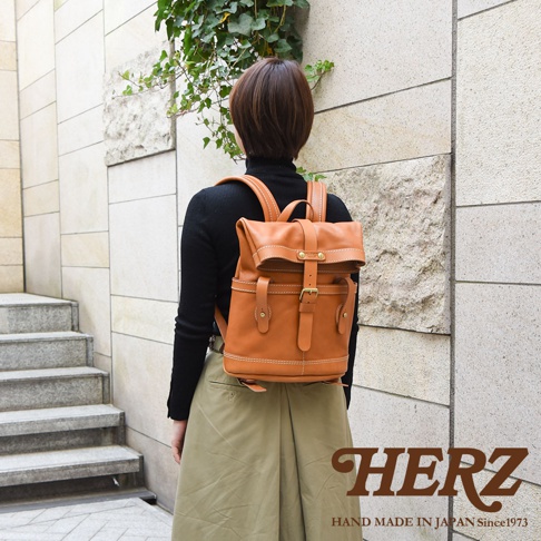 淺棕色皮革雙口袋折蓋後背包，配有可調式肩帶、金屬扣環和手工縫線細節。