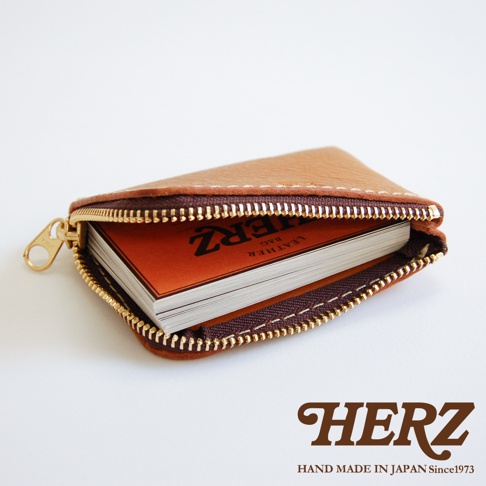 一張從側上方俯瞰的日本HERZ品牌手工皮革L型拉鍊收納夾，卡片夾採用柔軟的棕色Stare牛皮縫製，配備金色金屬拉鍊，袋口呈現L型開啟狀態，內層裝有多張印有HERZ品牌字樣的白色卡片，整體設計簡約俐落，展現細膩的手工皮革質感。