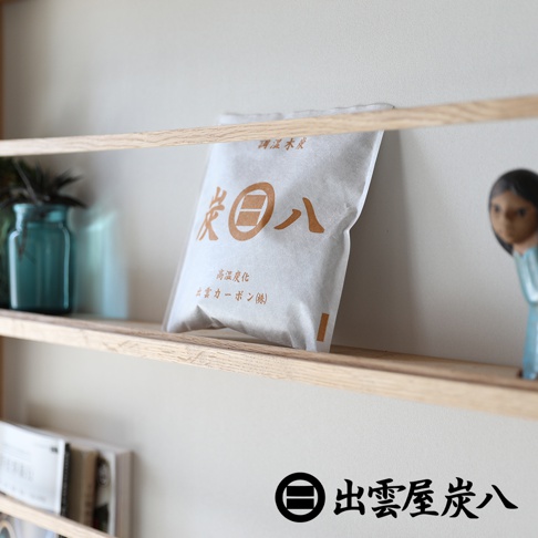木質層架上放置一包印有出雲屋炭八標誌的白色調濕木炭，旁邊擺放藍色玻璃瓶與精緻的人偶飾品，下方隱約可見書籍，整體呈現簡約的生活居家風格。