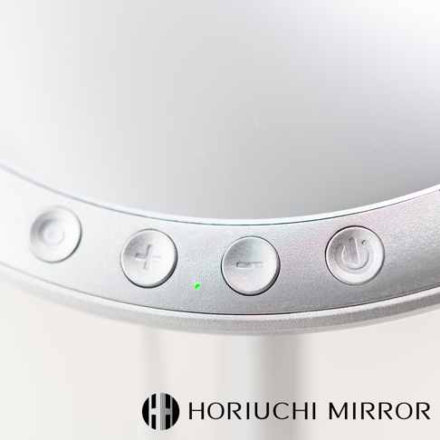銀色桌上型化妝鏡的局部特寫，顯示鏡面邊緣的控制按鈕，包括電源、加號、減號，以及一個綠色指示燈。