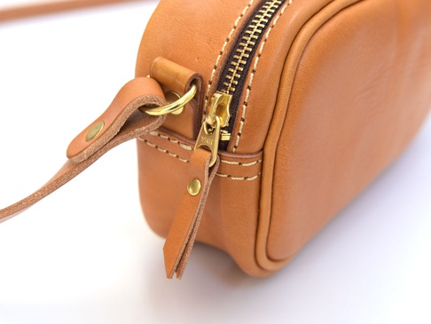 淺棕色皮革隨身單肩小包，金色 YKK 拉鍊，皮革肩帶連接處有金色卯釘和圓環。