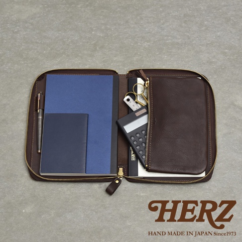 深棕色皮革製的拉鍊式記事本收納包，打開後可見左右兩側皆有收納空間，左側有藍色筆記本、深藍色小本子和一支筆，右側則有計算機、小剪刀和一個小型尺。收納包底部印有HERZ品牌字樣及HAND MADE IN JAPAN Since1973字樣。