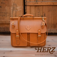 職人手作A-102皮革旅行包，採用經典棕色皮革，配有金色金屬釦件和可調節肩帶。包款設有頂部提把、翻蓋設計及雙前袋，縫線精緻，展現皮革的自然紋理。