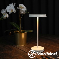 白色LED T-Light LOUNGE XL工型造型燈，頂部為圓盤形燈罩，燈身細長，底座為圓形，散發暖黃色光芒，旁邊擺放著一盆白色蝴蝶蘭，置於木質桌面。