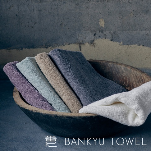 木碗中斜放著五條不同顏色的長纖浴巾，依序為淺紫、淺藍、米色、深藍，以及一條展開的白色浴巾，浴巾材質厚實蓬鬆。