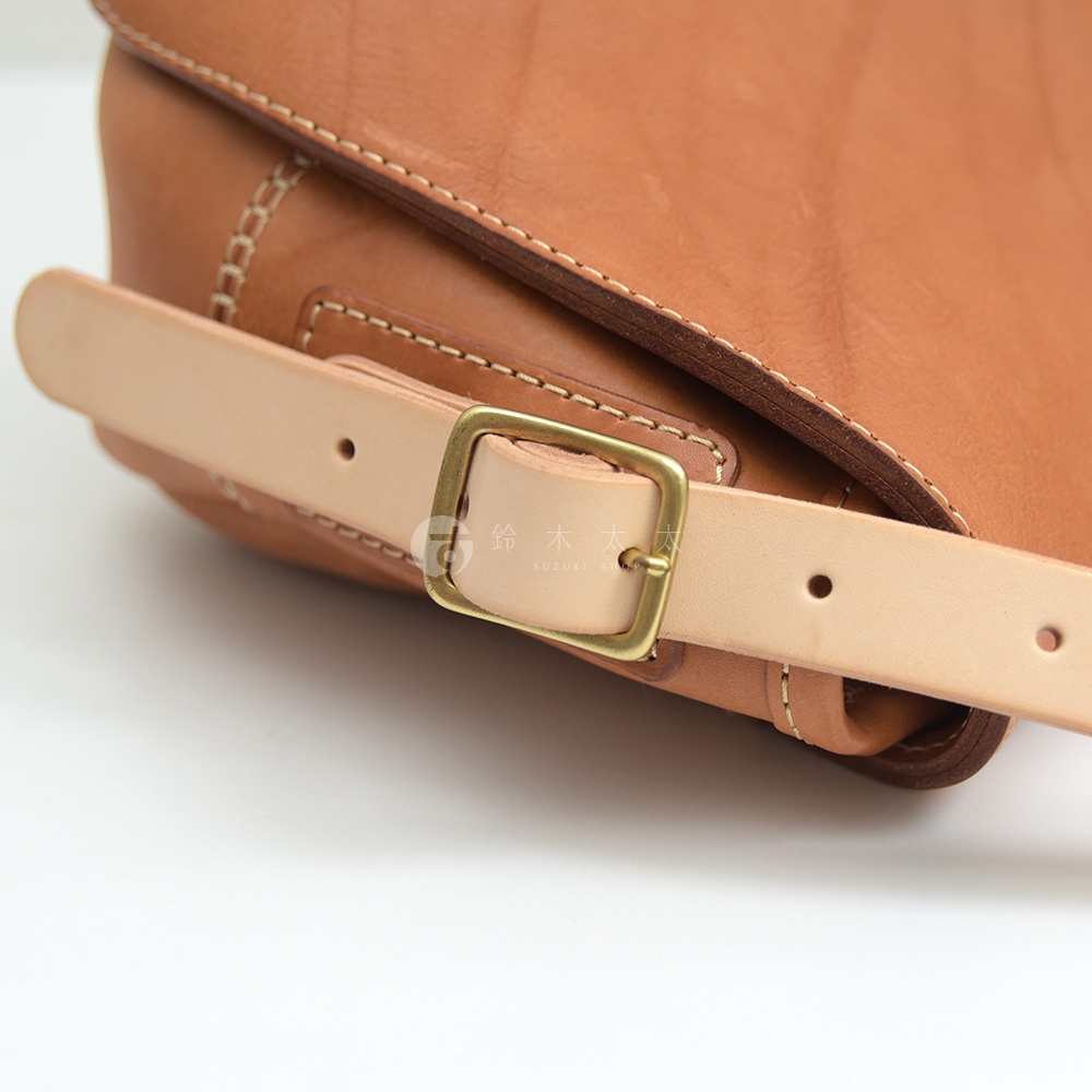 棕色皮革郵差包，配有淺棕色皮革繫帶和金色金屬扣環，縫線明顯。