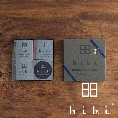 一張木紋背景上方擺放著 hibi 香氛火柴禮盒，左側為內裝三個小型火柴盒的開盒狀態，右側為外盒包裝並繫上一條點綴著小星星的藍色緞帶，下方則展示 hibi 的品牌標誌與文字。