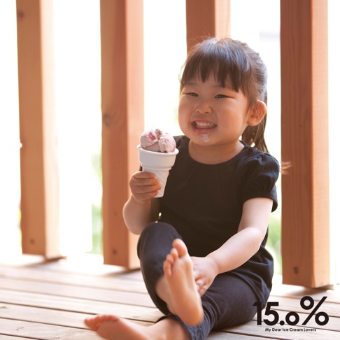 小女孩坐在木地板上，開心地拿著一個裝有粉紅色冰淇淋的白色甜筒杯，杯子下方是傳統的格紋甜筒。