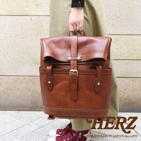 職人手作的棕色皮革雙口袋折蓋後背包，配有金屬釦環和皮帶束口，風格經典。
