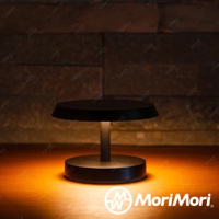 黑色金屬材質的LED T-Light LOUNGE工型造型檯燈，圓形燈罩下有簡潔的燈柱連接圓形底座，散發溫暖的黃色光芒，照亮桌面。