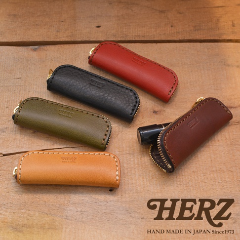 職人手作皮革印章收納袋 KZ-121，共展出五種顏色：紅、黑、軍綠、棕褐及淺棕。收納袋由皮革製成，配有拉鍊及金色吊飾，其中一個棕褐色的收納袋內裝有一支黑色筆狀物。