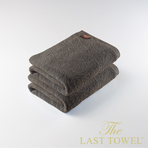 深棕色純棉柔浴巾堆疊，頂部一條繡有品牌標籤，質感厚實，整體呈暖色調。