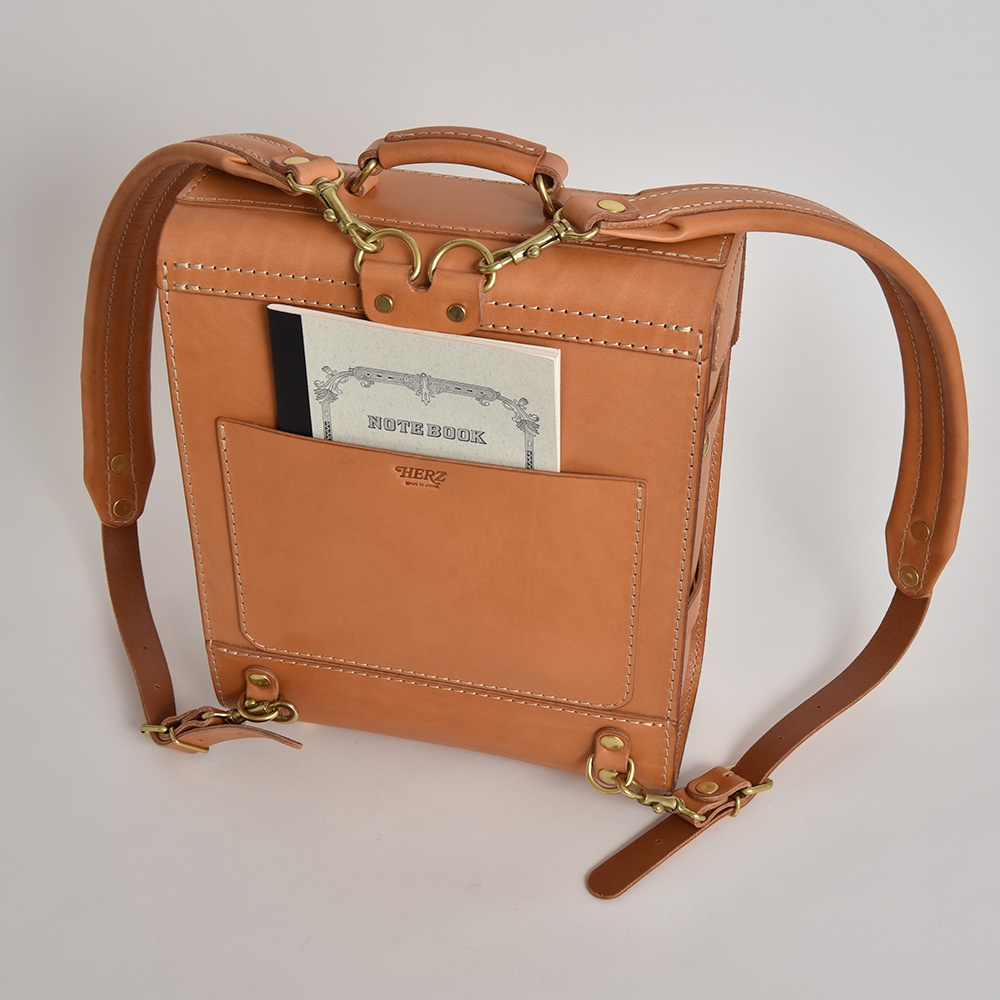 職人手作經典學院風後背包 R-100，採用全皮革材質，主體為淺棕色，配有可調節的肩帶、頂部提把，正面設有收納袋，袋中可見一本「NOTE BOOK」筆記本，整體設計簡潔實用。