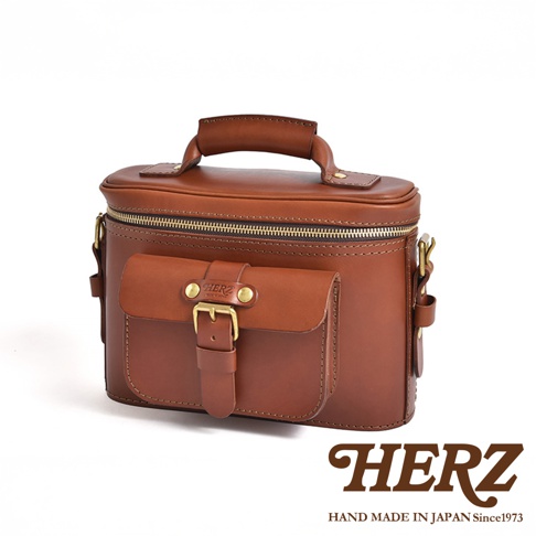 日本HERZ職人手作皮革厚實相機包，棕色皮革材質，帶有翻蓋口袋和皮帶扣，頂部有手提把，背景為白色。