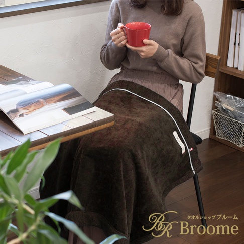 一個身穿咖啡色毛衣的女子，手裡拿著紅色杯子，坐在椅子上，膝蓋蓋著一條深棕色的毛毯，桌上放著一本雜誌，右下方有 Broome 的品牌標誌。