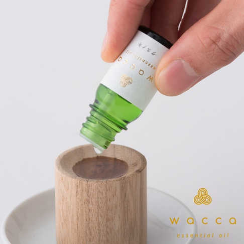 一滴綠色精油從標有「WACCA essential oil」字樣的白色標籤小玻璃瓶中滴落，置於一個圓形木製擴香石的凹槽中。