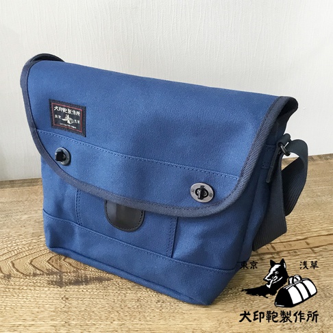 藍色純棉帆布雙扣郵差包，帶有黑色皮質裝飾和品牌標籤，斜挎式設計。