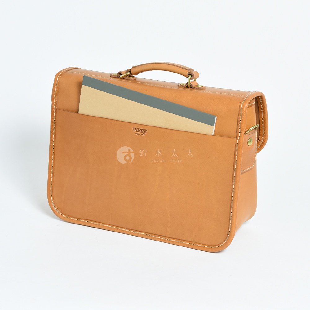 淺棕色皮革製作的兩用單肩包，包身方形，配有提把和可調節的側背帶，正面設有插袋，袋中放置一本筆記本。
