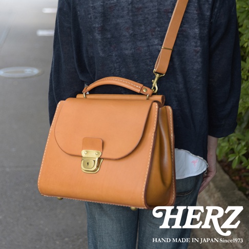HERZ 職人手作屋瓦風皮革兩用單肩包，以焦糖色硬皮革製成，搭配金色鎖扣與車縫線細節，展現日式簡約風格。