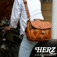 棕色皮革製成的鹿角扣繩郵差包，帶有縫線細節和可調節肩帶，由一位穿著白色襯衫和淺藍色牛仔褲的人攜帶，靠近摩托車。