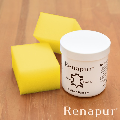 白色圓罐裝的 Renapur 皮革保養油，罐上印有品牌名稱、皮革圖標及「Extra Quality Leather Balsam」等字樣。旁邊擺放著兩塊黃色海綿，皆置於木紋背景上。