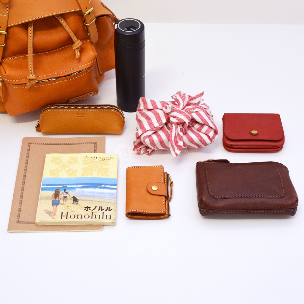 職人手作皮革復古風旅行後背包 R-65，搭配的配件包括：一個橘色皮革筆袋、一個黑色保溫杯、一塊紅白色條紋布包裹的物品、一個紅色皮革短夾、一本 Honolulu 旅行書、一個橘色皮革短夾，以及一個深棕色皮革長夾。