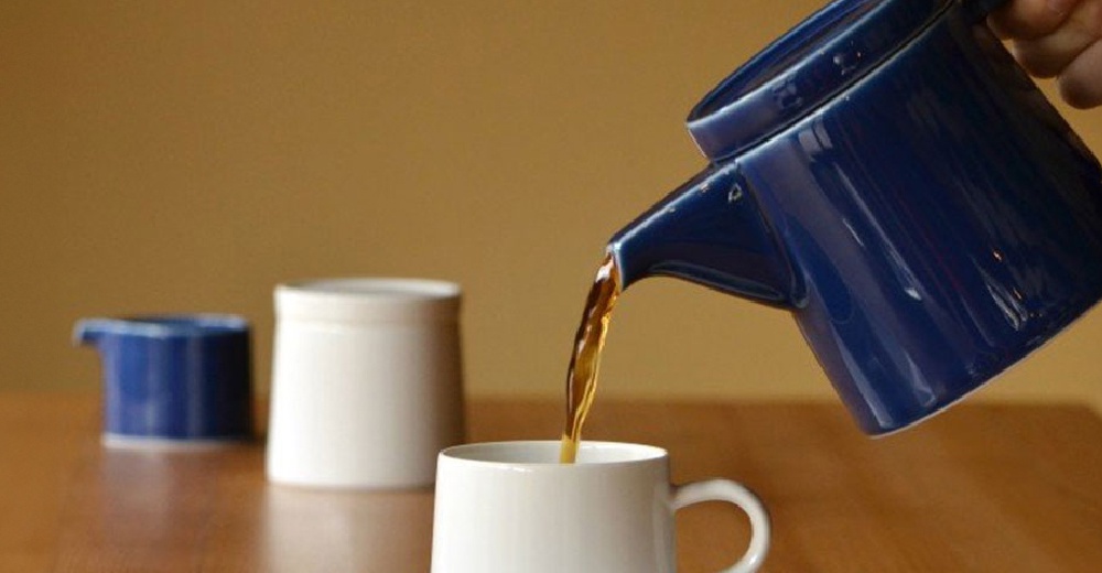 藍色陶瓷M型壺傾倒深棕色液體至白色馬克杯中，背景有另一藍色小壺和白色圓柱形容器。