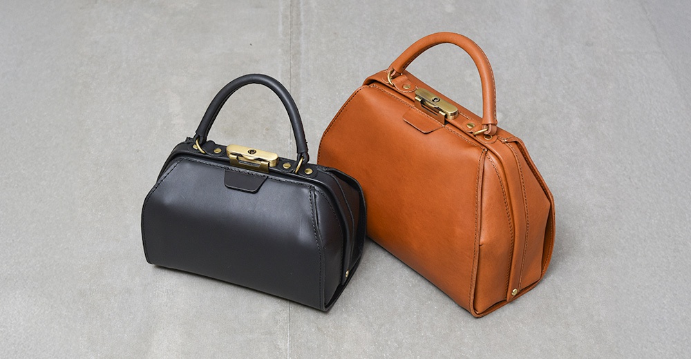 兩款職人手作皮革Organ兩用口金手提包，一為經典黑色，另一為溫潤的棕褐色，皆採用皮革材質，搭配金色金屬口金鎖扣和提把，呈現復古而精緻的風格。