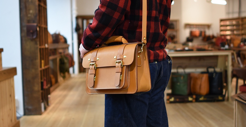 棕褐色皮革手提單肩包，具有兩個經典雙扣翻蓋和金屬細節。包款配有可拆卸的肩帶，由兩人展示。