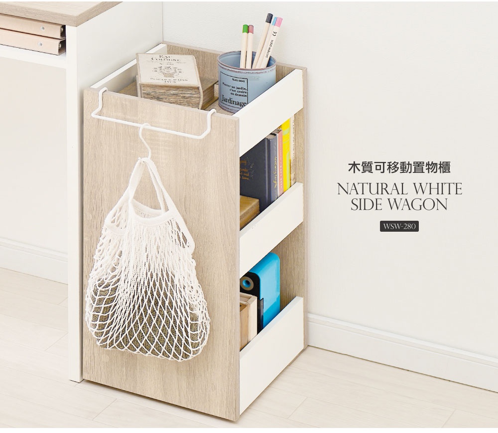 木質可移動置物櫃，側邊附有掛架，掛著一個白色網狀購物袋。置物櫃為淺木紋色，搭配白色邊框，內有三層收納空間，擺放了書籍、筆筒及藍色削筆器。
