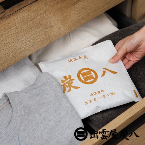 一隻手將印有出雲屋炭八品牌標誌與文字的白色方形調濕木炭包，放置在收納抽屜內衣物與毛巾之間，呈現居家生活收納場景。