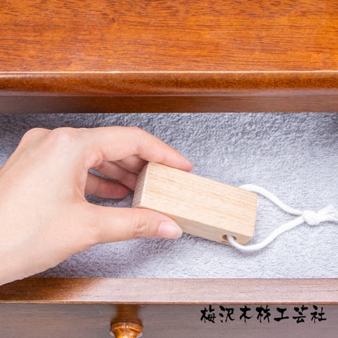 一隻手拿著一塊淺木色、長方形的樟木條，上面繫著一條白色細繩。木條置於灰色毛巾上，背景為木質抽屜。
