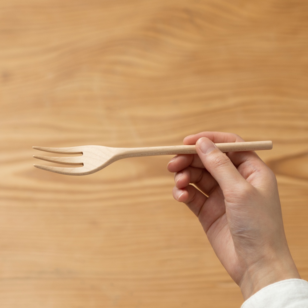一隻手拿著一支淺色楓木製的餐叉，餐叉有三個叉齒，背景為木紋桌面。