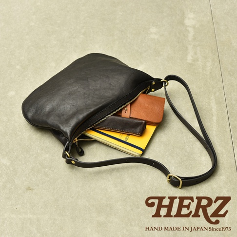 黑色職人手作皮革Organ單肩半月包，包身半月形，開口處有金色拉鍊，內側可見黃色筆記本、棕色錢包及深棕色皮夾。