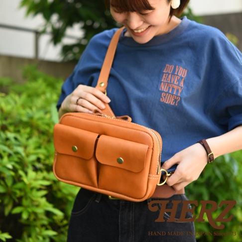 一款棕色皮革製成的雙口袋斜背包，中間有拉鍊開口，兩個前袋均有金屬釦裝飾，由一位女性展示。