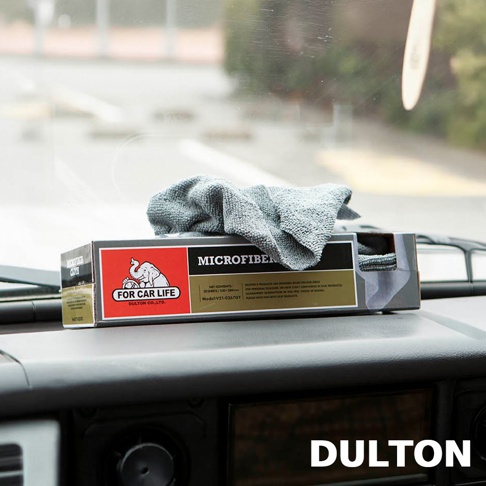 一盒 DULTON 品牌的 FOR CAR LIFE 超細纖維擦拭布放置在汽車儀表板上，盒身印有可愛的象形圖標，頂部露出一塊灰色的超細纖維布，背景為模糊的車外景觀。