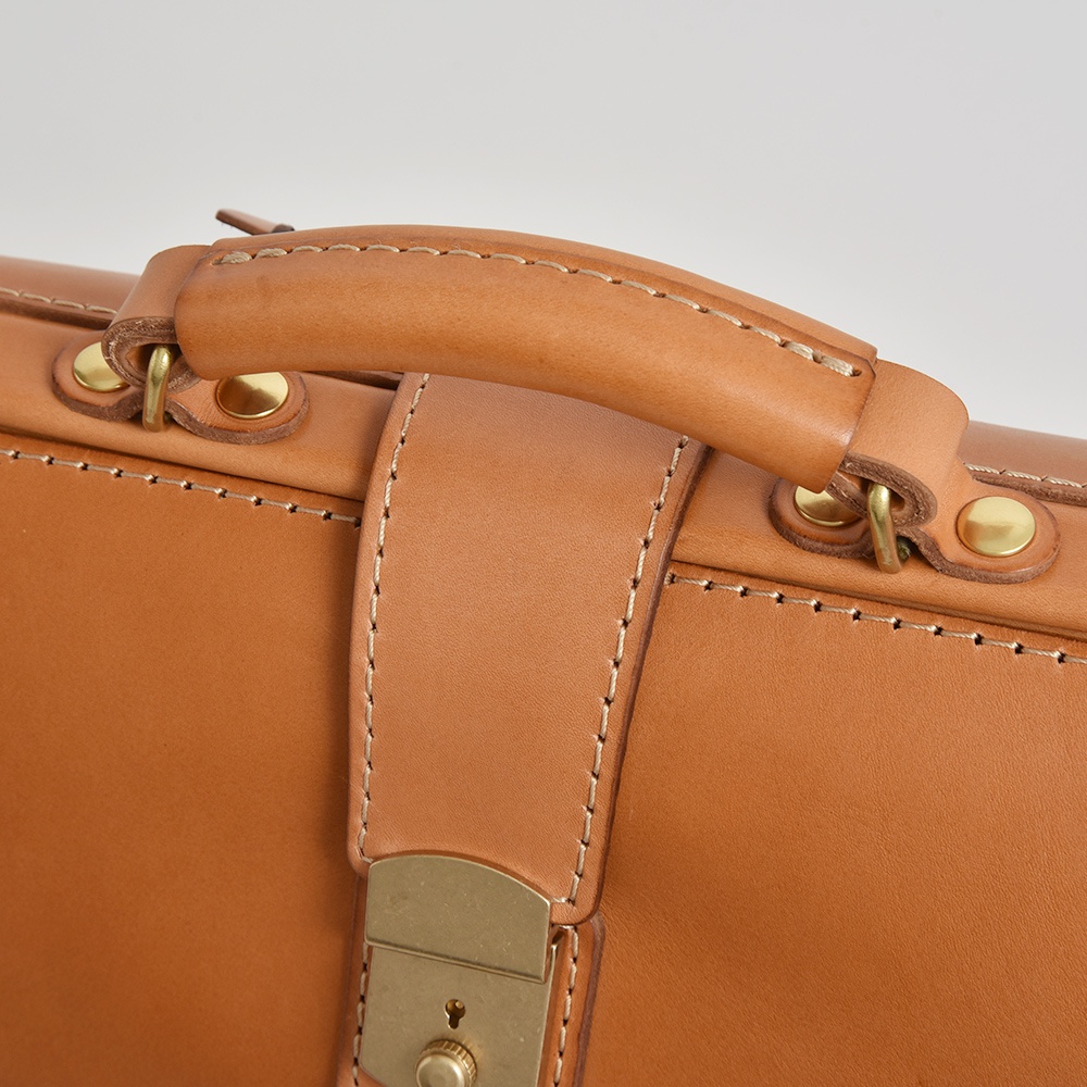 焦糖色皮革公事包特寫，展示手提把、縫線細節、金色鎖扣及鉚釘。