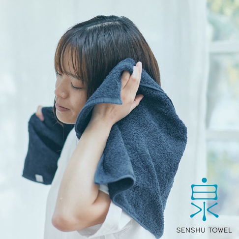 深藍色 SOLID 細柔蓬韌緞澤長纖浴巾，質地蓬鬆，觸感細膩，正在被一位女性用於擦拭臉部。
