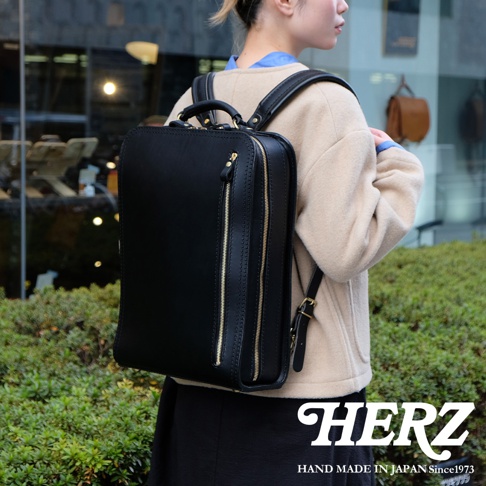 黑色皮革商務電腦後背包，具有方形輪廓、頂部提把、兩個拉鍊前袋和可調節肩帶，由職人手工製作。