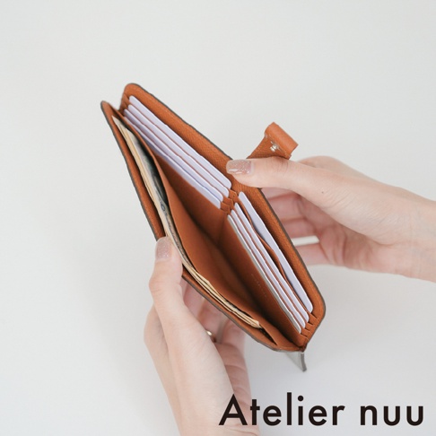 一雙白皙的手正溫柔地撐開 Atelier nuu 品牌駝色皮革雙折長夾，展示內部多層卡片收納槽與存放紙鈔的夾層，整體呈現極簡質感與細膩皮革紋理。