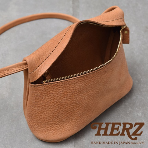 HERZ 品牌標誌，下方為 HAND MADE IN JAPAN Since 1973 字樣，整體為一個 tan 色皮革製成的斜背包，包身紋理細緻，拉鍊為金色，包包開口朝上，呈現未使用的狀態，背景為灰色水泥地。