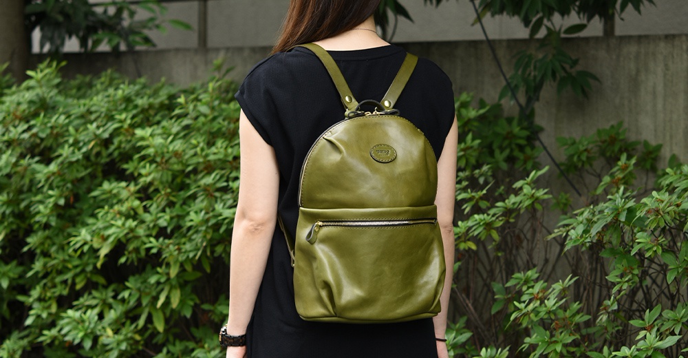職人手作皮革大開口圓角後背包，橄欖綠色，帶有拉鍊前袋和圓形壓印品牌標誌。